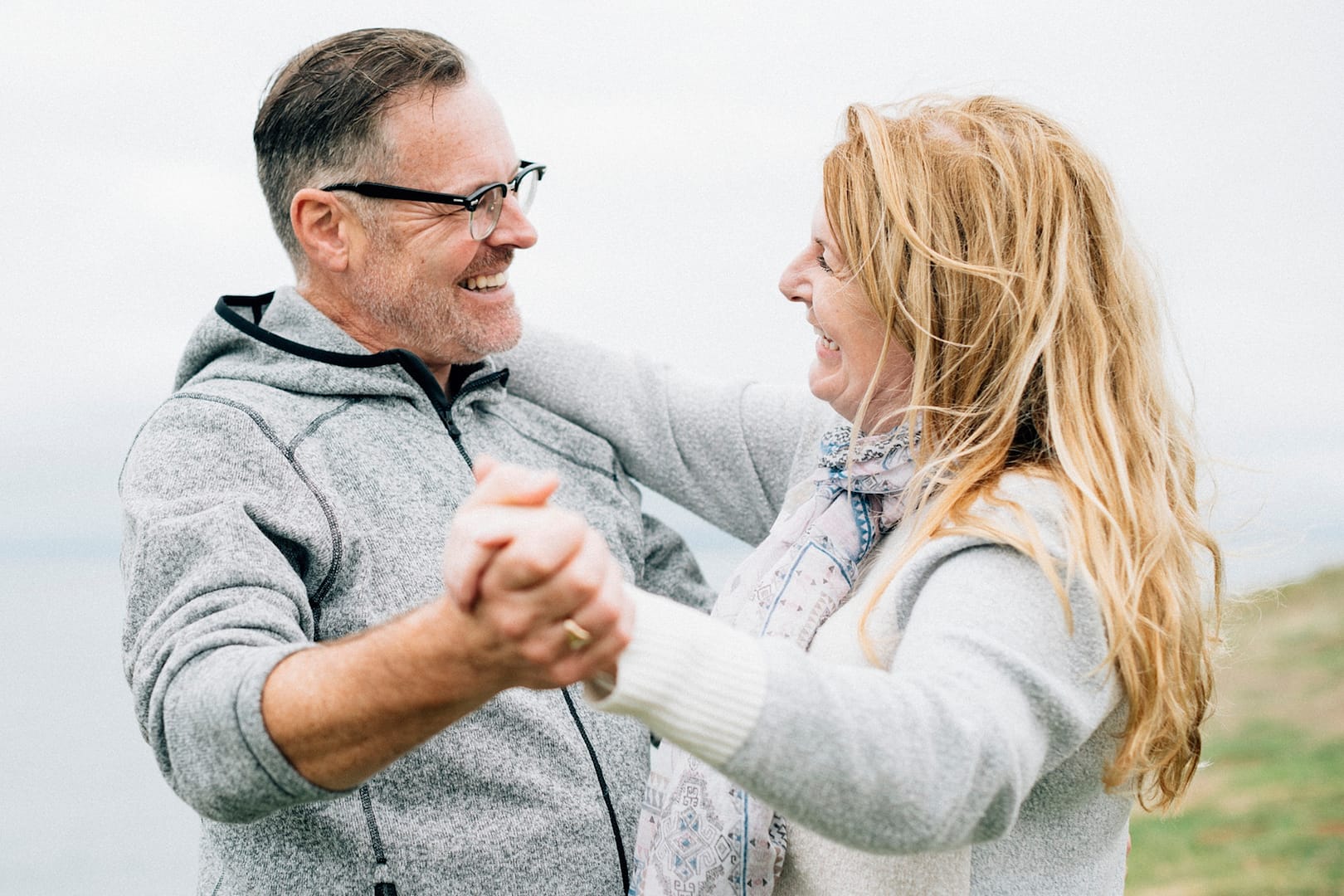 Happy middle-aged couple dancing outdoors on a breezy day, sharing a joyful moment of connection and love, with a scenic natural background.