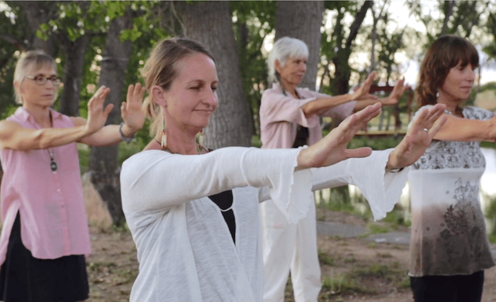 Group of women practicing Qigong outdoors in a peaceful wooded area, moving their arms gracefully in unison, symbolizing mindfulness, harmony, and connection with nature.