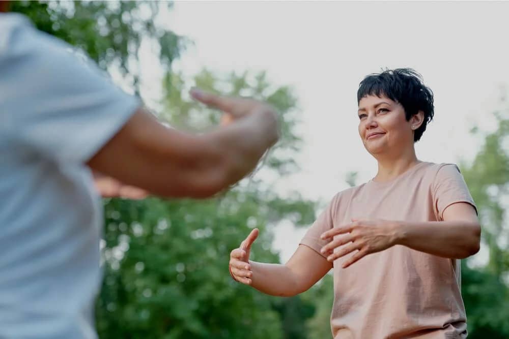 Two individuals practicing Qigong outdoors, focusing on gentle and harmonious hand movements, surrounded by a tranquil natural setting with lush greenery.