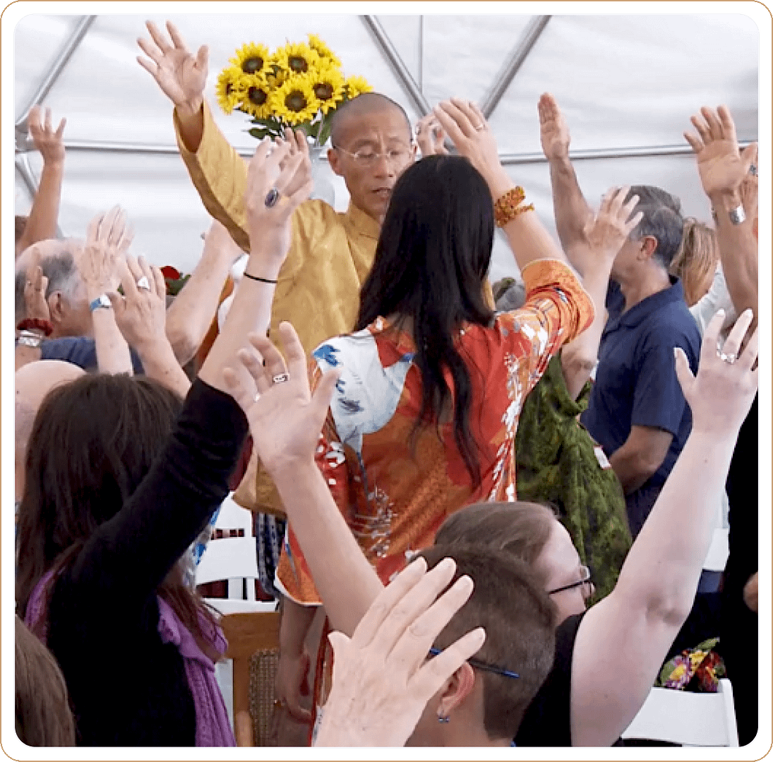 Master Mingtong Gu leading a vibrant Qigong session in a dome setting, surrounded by participants raising their hands, with bright sunflowers in the background adding a touch of nature and warmth.