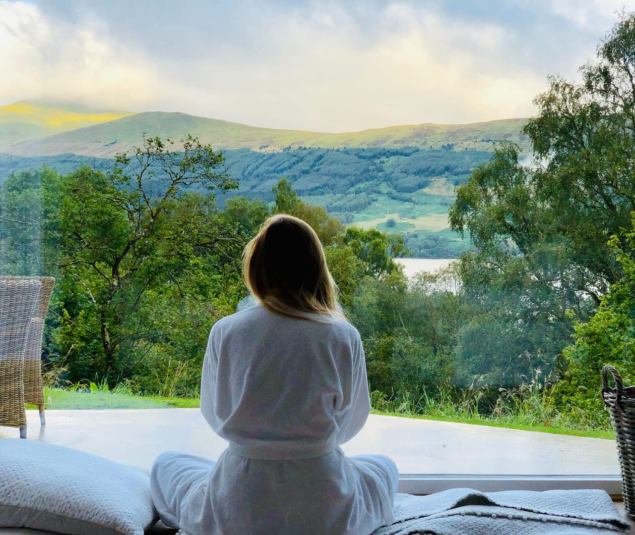 A woman sitting on a bed practicing Qigong, looking out a window.