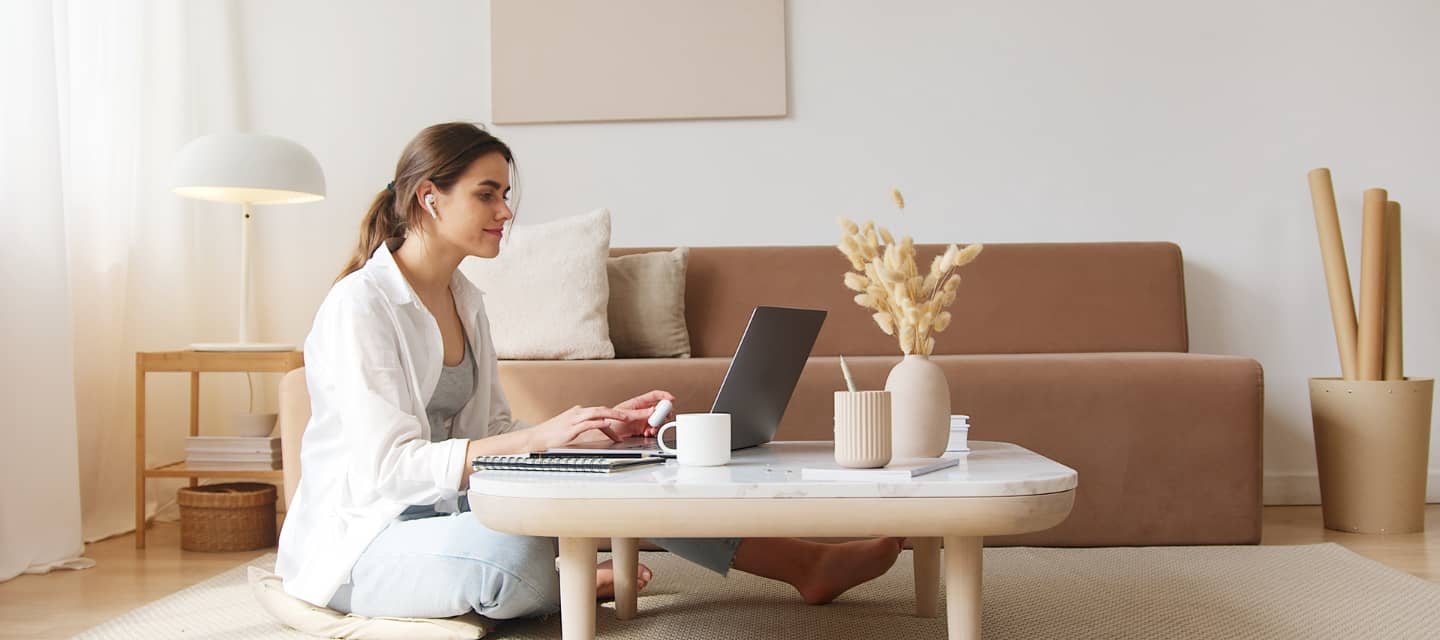 A young woman sitting cross-legged on the floor in a cozy, minimalist living room, working on a laptop with earphones, surrounded by a notebook, coffee mug, and a vase with dried flowers, symbolizing focus, comfort, and remote learning