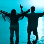 Two individuals stand waist-deep in calm water with their backs to the camera, arms outstretched in a gesture of freedom and joy. The figure on the left holds a sheer scarf billowing in the breeze, while the figure on the right raises one arm higher. The scene is bathed in a vibrant blue hue, evoking a sense of serenity, unity, and connection to nature.