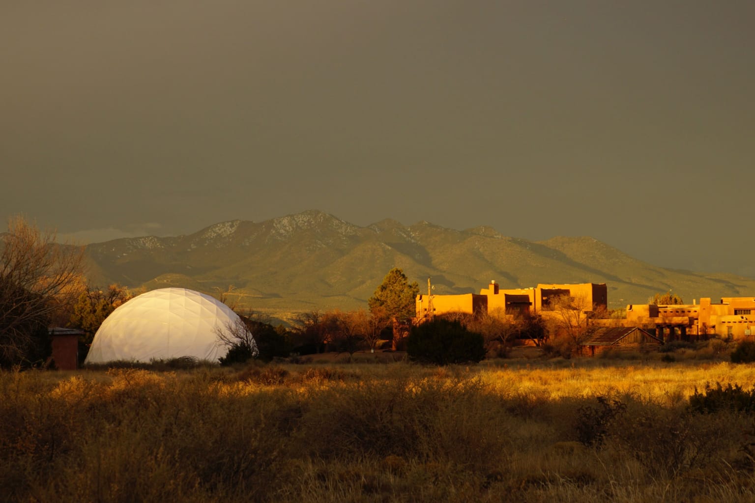 Scenic view of the Chi Center Dome with golden adobe structures in the foreground, set against a majestic mountain backdrop under a dramatic sky