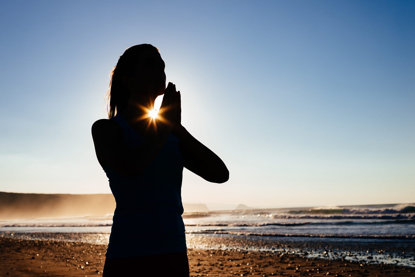 Silhouette of a woman in a meditative prayer pose with hands pressed together, framed by the rising sun shining through her heart space, against a clear blue sky.