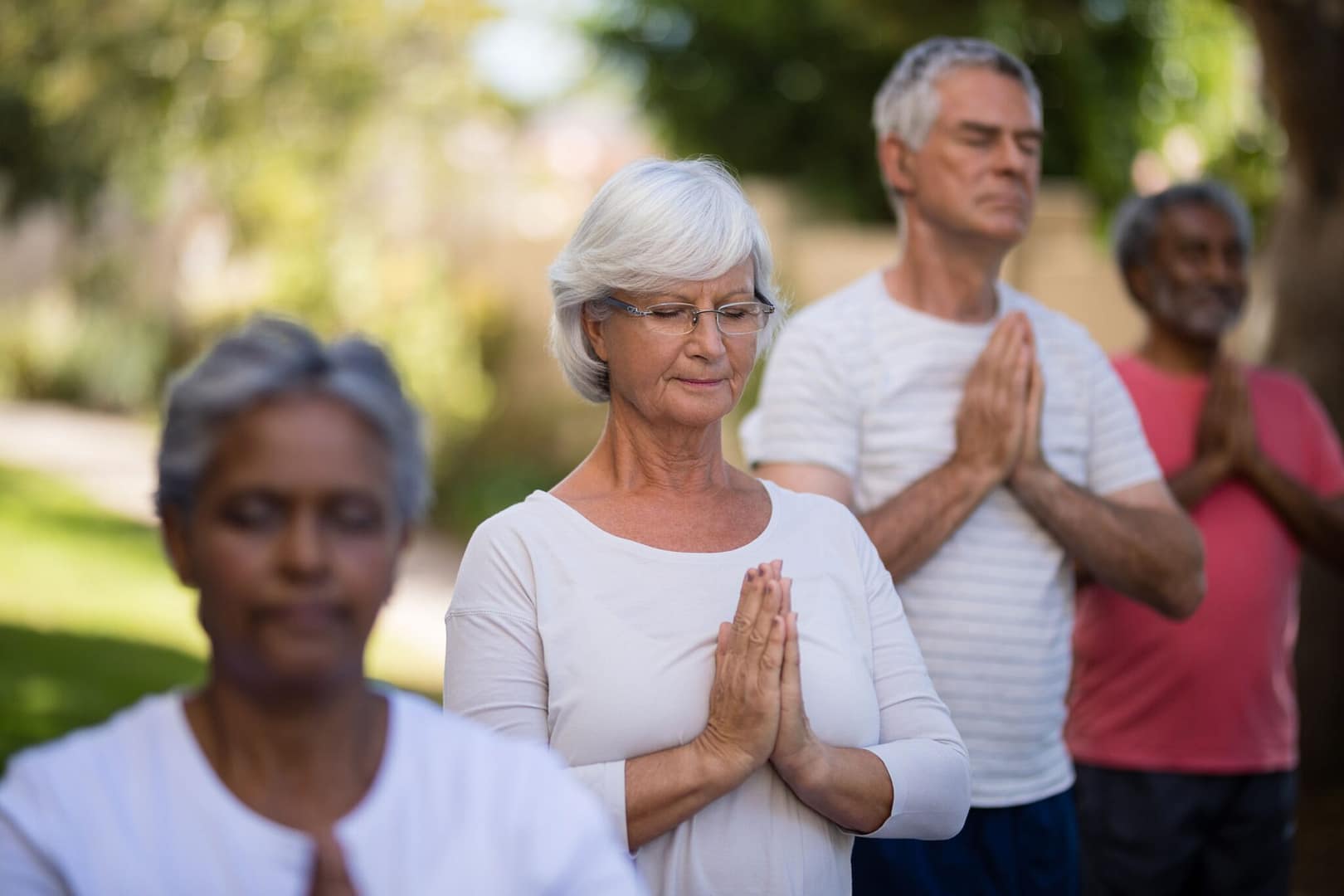 A group of people meditating outdoors, standing with hands in a prayer position and eyes closed. The serene expressions and natural setting create a peaceful atmosphere of mindfulness and connection.
