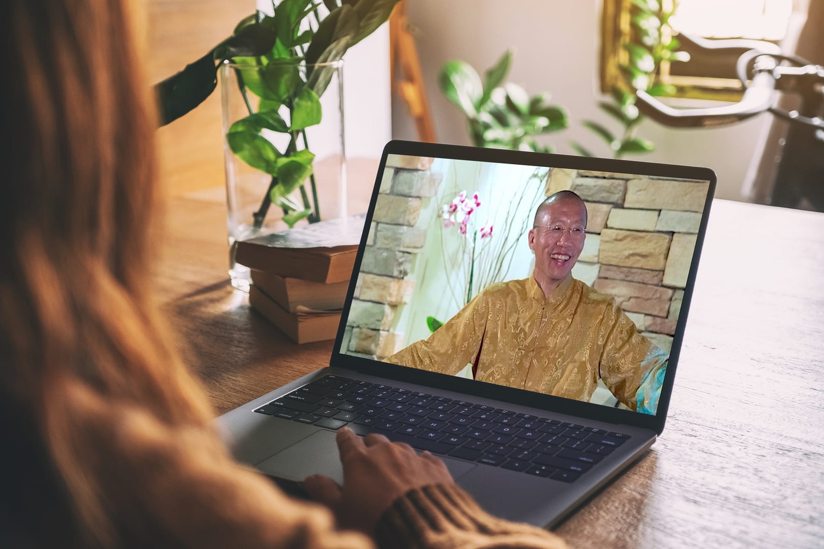 Person watching a video of Qigong Master Mingtong Gu in a golden robe, smiling against a stone wall backdrop, with plants and books on a wooden desk, representing online wellness learning.