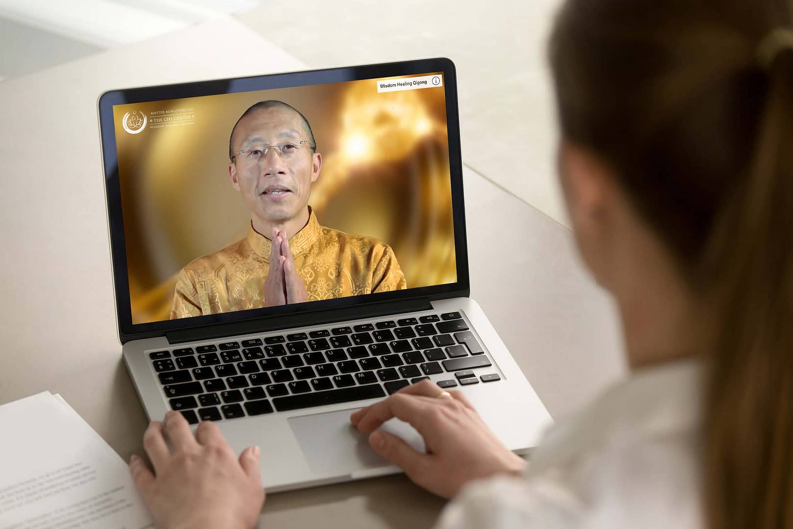 Student participating in an online Wisdom Healing Qigong session with Master Mingtong Gu displayed on a laptop screen, wearing a golden traditional outfit, set against a warm glowing background.