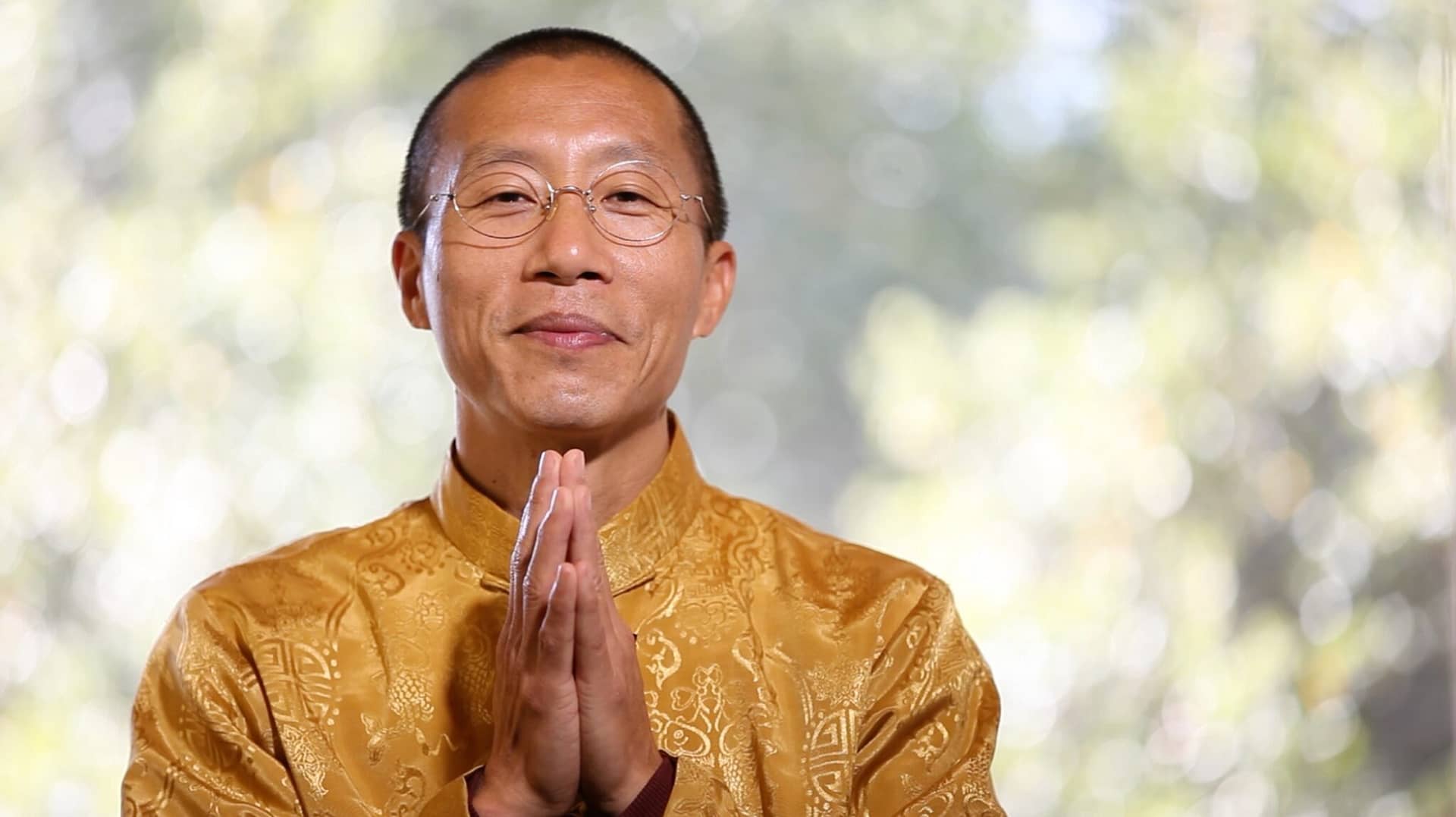 Master Mingtong Gu in a serene pose, wearing a golden traditional garment, offering a gesture of gratitude and mindfulness against a softly blurred natural background.