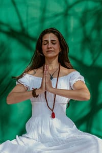 Serene woman meditating with hands in prayer position, wearing a white dress and mala beads, set against a vibrant green background with natural shadow patterns.