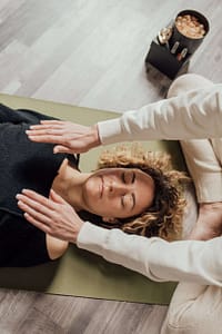 A woman with curly blonde hair is lying down on a green mat with her eyes closed, appearing relaxed. Another person, only visible from the arms, is gently placing their hands on the woman's chest and shoulder area. The setting seems to be a calming wellness or therapy session. On the floor beside them, there's a box with some items, including what appears to be crystals. The scene is set on a light-toned wooden floor.