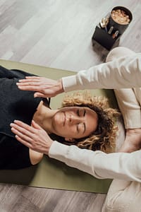 A woman with curly blonde hair is lying down on a green mat with her eyes closed, appearing relaxed. Another person, only visible from the arms, is gently placing their hands on the woman's chest and shoulder area. The setting seems to be a calming wellness or therapy session. On the floor beside them, there's a box with some items, including what appears to be crystals. The scene is set on a light-toned wooden floor.