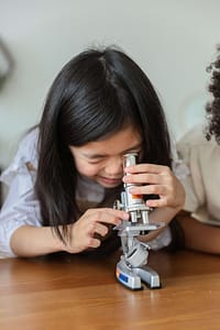 A young girl with dark hair is intently looking through a microscope placed on a wooden table. She delicately adjusts the equipment with her fingers. The microscope has a silver and gray base with a red and orange color tube. A partial view of another person with curly hair is seen to the right of the image. The background is soft-focused and neutral-toned.