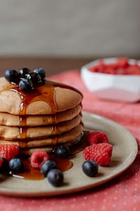 A stack of fluffy pancakes topped with fresh blueberries, drizzled with maple syrup, and surrounded by vibrant raspberries, served on a rustic beige plate with a bowl of additional raspberries in the background