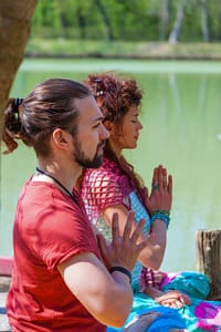 Two individuals meditating peacefully outdoors near a calm lake, with their hands in a prayer position, surrounded by vibrant greenery and sunlight.
