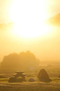 Golden sunrise over a peaceful campsite with a picnic table, orange tent, and scattered rocks, surrounded by mist and warm morning light in nature.