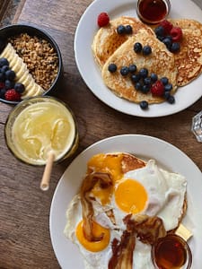 A delicious breakfast spread featuring fluffy pancakes topped with fresh blueberries, raspberries, and syrup, sunny-side-up eggs with crispy bacon, and a side of granola, banana, and berries, accompanied by a refreshing glass of juice.