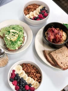 A vibrant breakfast display featuring avocado toast garnished with lemon and chives, yogurt bowls topped with granola, bananas, blueberries, and raspberries, a bowl of creamy oats with crispy bacon, and slices of fresh sourdough bread.