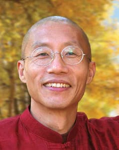 A portrait of Master Mingtong Gu, a man with a warm smile, wearing round glasses and a red traditional top. The background features vibrant autumn colors, with golden-yellow leaves creating a peaceful and radiant atmosphere.