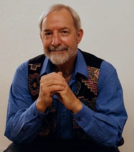 Portrait of Len Schreiner, dressed in a blue shirt and patterned vest, seated with hands clasped, exuding wisdom and approachability.