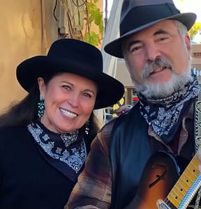 Members of Roadside Distraction, a duo dressed in Western-inspired attire with black hats, turquoise earrings, and patterned bandanas, smiling warmly. One holds a guitar, adding a musical touch to the friendly and inviting atmosphere.
