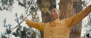 Master Mingtong Gu practicing Qigong outdoors with arms open wide, wearing a golden traditional Chinese jacket, standing peacefully in front of tall trees—symbolizing healing, energy flow, and connection with nature.