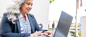 A smiling woman with long silver hair working on a laptop outdoors, showcasing focus, productivity, and professional engagement in a bright, urban setting.