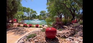 Serene garden setting on Magical Sunday, featuring a vibrant red water fountain surrounded by smooth river stones, a tranquil pond with a central fountain, and bright red Adirondack chairs under the shade of lush trees, creating a peaceful retreat atmosphere.