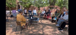 A serene gathering at Magical Sunday, with participants seated in a circle outdoors, attentively listening to a speaker dressed in a golden shirt. The group is surrounded by trees and natural beauty, creating a peaceful and reflective atmosphere.