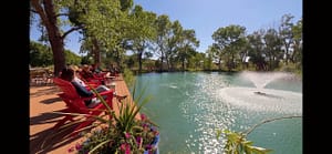 Attendees relax in vibrant red Adirondack chairs by a peaceful turquoise pond on Magical Sunday, with sunlight sparkling on the water's surface and a fountain gently creating ripples, surrounded by lush green trees.