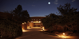 Night view of the entrance to the Kiva at The Chi Center, illuminated under a clear sky with a full moon. The path to the Kiva is lined with glowing lanterns, casting a warm light on the surrounding dense, green shrubbery. This tranquil setting invites a peaceful environment suitable for Wisdom Healing Qigong practice and spiritual rejuvenation.