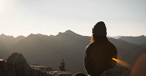 A person in contemplation, sitting on a rocky ledge overlooking a vast range of mountains during sunset, embodying life mastery and the serenity of Qigong.