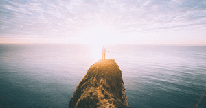 A solitary person stands on a narrow cliff above a vast ocean, bathed in the warm light of the setting sun, symbolizing contemplation and connection with nature.
