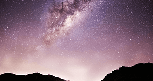 Starry night sky over a silhouette of mountain ranges with the Milky Way galaxy prominently displayed in shades of pink and purple, illuminating countless stars scattered across the heavens.