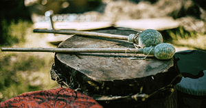 Close-up of a traditional drum and mallets resting on an outdoor setting, symbolizing the ancient practice of sound healing. The weathered drum and handcrafted mallets are surrounded by a natural backdrop, reflecting the principles of Hun Yuan Chi and Wisdom Healing Qigong, emphasizing the connection between rhythm and source energy.