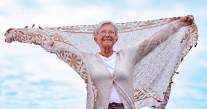 Elderly Caucasian woman joyfully extends a patterned shawl against a soft blue sky, embodying freedom and happiness. Her radiant smile and uplifting pose reflect her commitment to self-love and well-being, enhanced by practices like Wisdom Healing Qigong that foster health and spiritual connection.