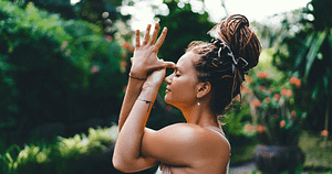 A young woman with dreadlocks tied up high practices a Wisdom Healing Qigong gesture, closing her eyes in a serene garden setting. Her posture and the peaceful environment emphasize a connection to self-love, spirituality, health, and well-being, reflecting a deep engagement with her inner self amidst vibrant, green foliage.