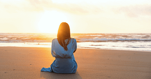 A woman in a blue robe sits peacefully on a sandy beach at sunset, contemplating the ocean. This tranquil scene embodies the practices of Wisdom Healing Qigong and principles of self-love, spirituality, health, and well-being, as the golden sunlight bathes her in a warm glow, enhancing the serene atmosphere.