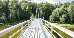 A modern white suspension bridge spanning over a tranquil river, surrounded by lush green forests. The bridge, bathed in sunlight, represents a journey towards self-love and spiritual well-being, reflecting the principles of Wisdom Healing Qigong which emphasize harmony with nature and personal health.
