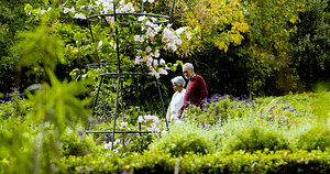 An elderly couple walking hand in hand through a lush, vibrant garden filled with blooming flowers, rich greenery, and hints of autumn colors. This image embodies the theme of The Healing Power of Nature: A Holistic Exploration, symbolizing the joy, calm, and rejuvenating effect of being immersed in natural surroundings, as well as the healing presence of shared moments in a tranquil setting.