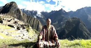 Master Mingtong Gu, wearing a colorful, traditional woven garment, sits cross-legged in meditation on a grassy hill overlooking the ancient stone ruins of Machu Picchu.