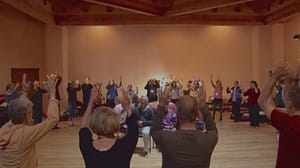 A group of people in a spacious, warm-toned room practicing a communal movement exercise, with their arms raised overhead in synchrony. The participants appear engaged and energized, creating a sense of unity and shared purpose in this uplifting practice session.