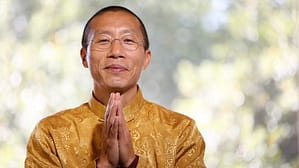 Master Mingtong Gu in a serene pose, wearing a golden traditional garment, offering a gesture of gratitude and mindfulness against a softly blurred natural background.