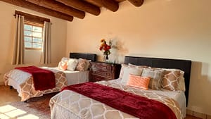 Bright and cozy bedroom featuring two beds with plush, patterned bedding, red throws, and decorative pillows. Natural light streams through the windows, complementing the rustic wooden beams and soft neutral tones of the space. A vase of vibrant flowers decorates the dresser, adding warmth to the room's serene ambiance.