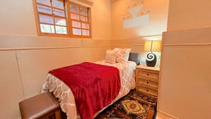 Serenity Center Room #5 - PURITY at the Southwest Sanctuary. Cozy single room with a warm red throw blanket, soft beige bedding, natural wood nightstand, and a decorative wall mural. Relaxing and peaceful retreat space for rejuvenation.
