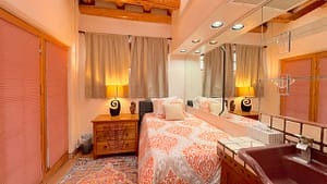 Serenity Center Room with single bed, bright orange and white bedding, wooden nightstand, decorative lamp, and built-in mirror wall. Features a maroon sink and cozy Southwestern design with warm wooden ceiling beams and neutral curtains.