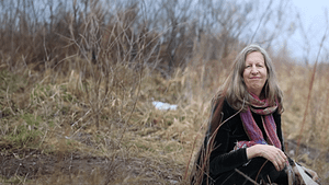 Debora Seidman sits peacefully amidst a natural, earthy setting, surrounded by dry grasses and twigs. She has a gentle, contemplative smile, draped in a warm scarf that adds a touch of color to the muted background, embodying a sense of calm and introspection for her Wisdom Healing Qigong testimonial."