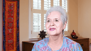 Bianca shares her Wisdom Healing Qigong journey, her face radiating calmness and wisdom. She is seated in a warm, inviting space adorned with colorful wall hangings and natural light streaming through nearby windows.