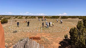 A diverse group of people engaging in a Wisdom Healing Qigong practice within a large stone labyrinth set in a vast, open field under a clear blue sky. The labyrinth, constructed from neatly arranged white stones, spirals into a center point, surrounded by sparse desert vegetation and distant hills, reflecting the serene environment of The Chi Center. Participants of various ages and ethnicities walk along the path in contemplative silence, enhancing the spiritual and healing atmosphere of the setting.
