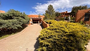 A sunlit pathway lined with vibrant green juniper and golden shrubs leads to The Chi Center, where people enjoy the peaceful outdoor setting under a clear blue sky.
